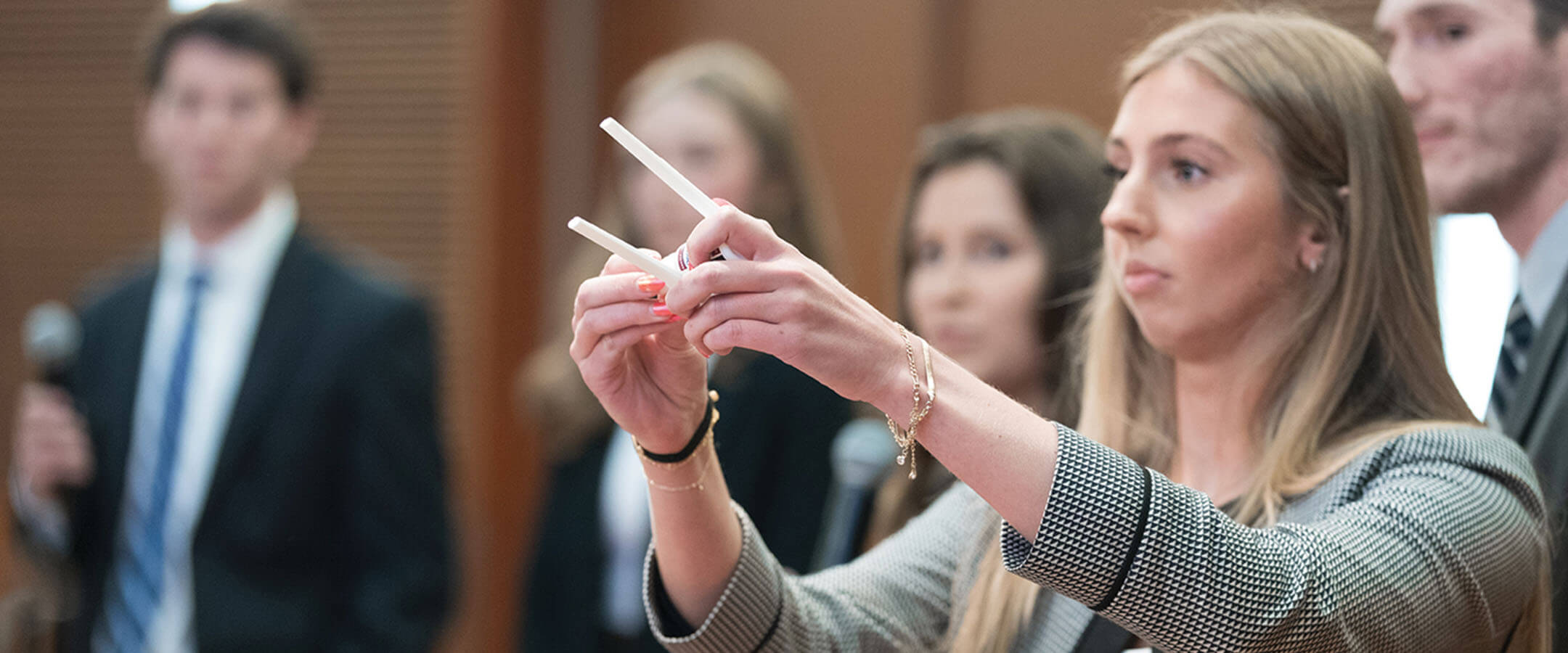 A young woman with long blonde hair presents a small, white, two-pronged device. She is wearing a black-and-white checked blazer over a white shirt. There is a silver bracelet on her right wrist and a black hairband on her left wrist. Her arms are outstretched to present the device to someone she is looking at out of frame. There are people out of focus behind her, also wearing formal business attire, watching her present the device.