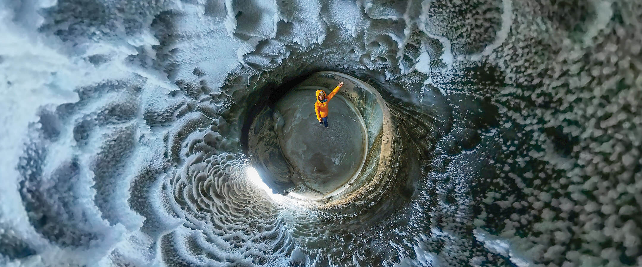 A woman is photographed from above and through a hole in some ice. The ice is dark white white whorls. The woman appears very small and far away, and she is wearing a yellow coat with the hood up and dark pants. She has her right arm raised in the air.