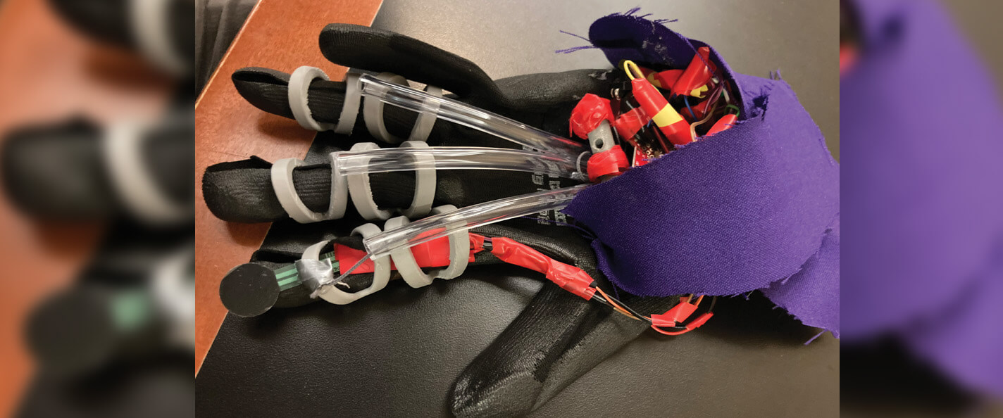 A black glove is displayed on a black mat. There are four grey silicone rings on the index, middle, and ring fingers. There is a red strap running up the index finger under the grey rings. There are three clear tubes running over the three middle fingers. There is purple fabric at the opening of the glove.