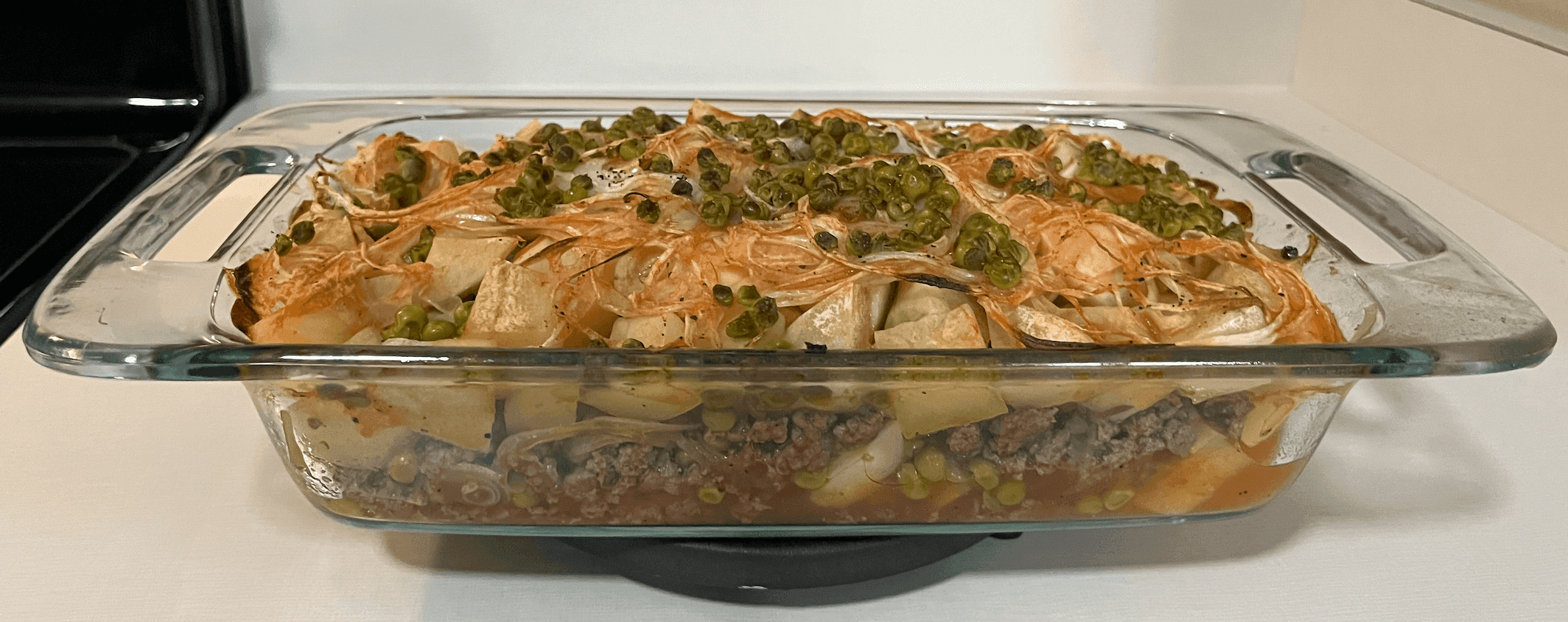 Clear baking dish with meat, potatoes, peas, and onions.