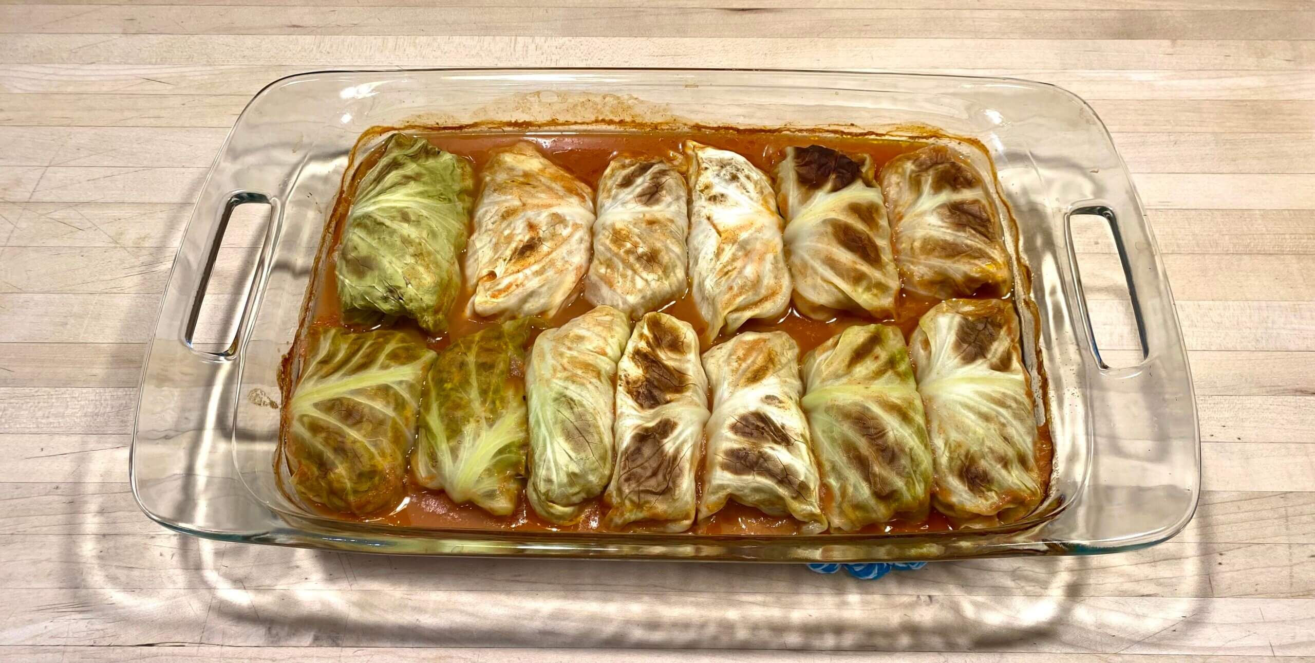 A glass baking dish sits atop a light-wash wood surface. The dish contains thirteen stuffed and rolled cabbage leaves submerged in a brown gravy.