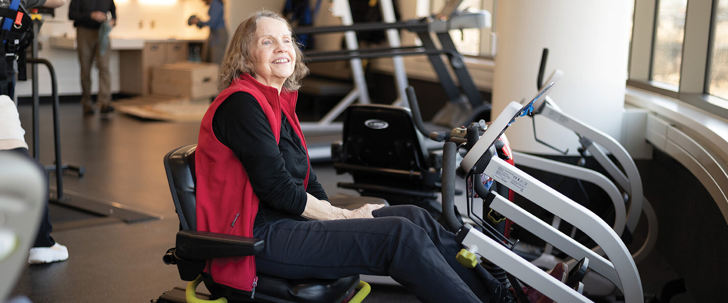 A woman sits on a seated exercise machine. She is wearing black pants and a black long-sleeve shirt and a red vest. She has short brown hair and is lit from the right by natural light. 