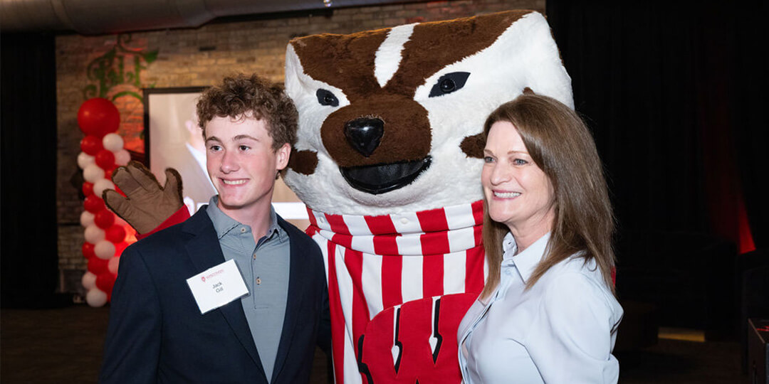 Three individuals pose for a photo, with the University of Wisconsin-Madison mascot Bucky Badger in the center; the person on the left wears a dark suit jacket with a name tag, and the person on the right wears a light-colored blouse. Red and white balloon columns are in the background.