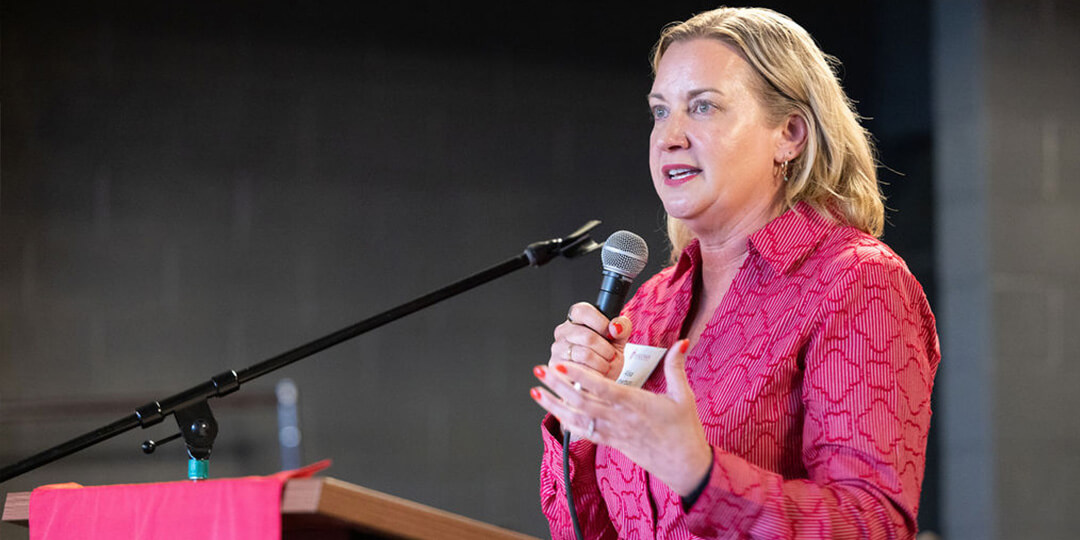Alisa Robertson, wearing a red shirt, speaks into a microphone at a podium.