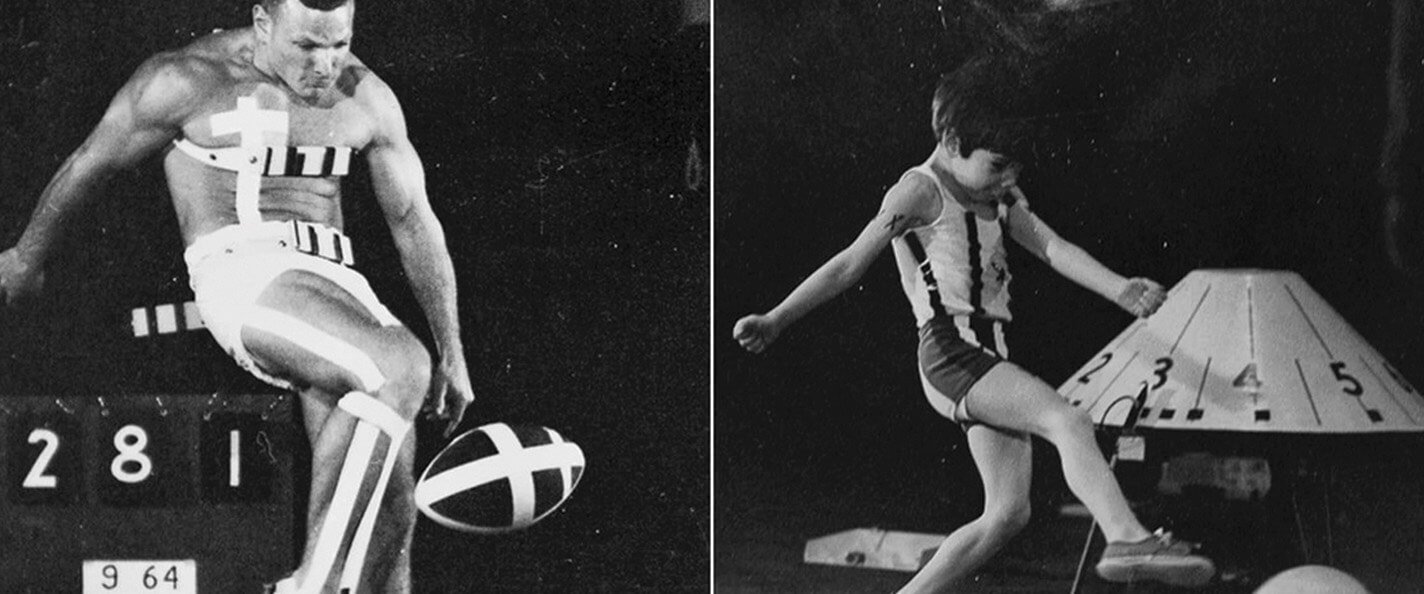 A black-and-white image of a study on the movement of a kick. On the left, a grown man wears white shorts and sneakers and kicks a football, his left leg raised and bent at the knee, his arms back, his shoulders hunched, and his head down. His left leg and left side-body feature white tape to highlight the position and shape of his body through the motion. He is standing on a dark floor and set against a dark background.