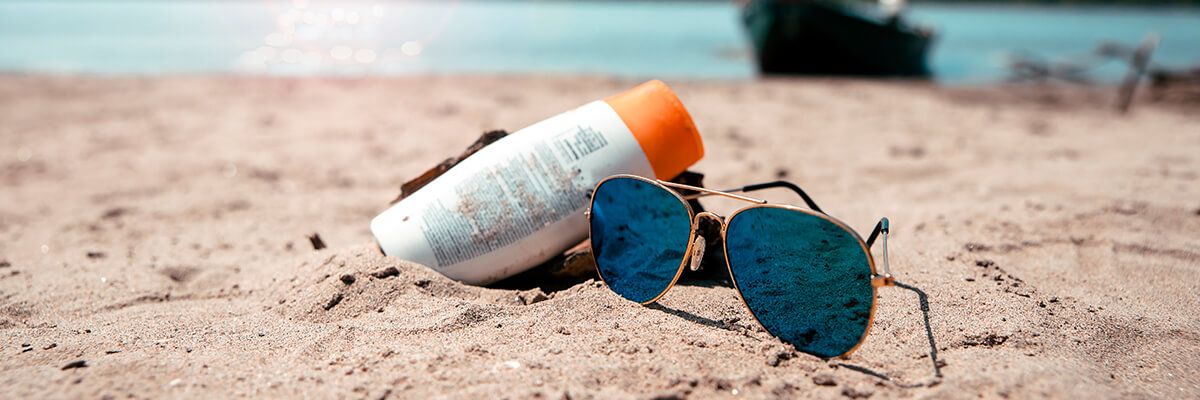 A bottle of sunscreen leans on a pair of aviator sunglasses on a beach. There is turquoise water visible in the background.