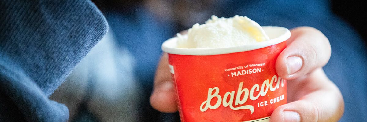 A close-up image of a hand holding a red cup of vanilla ice cream. The cup says "Babcock" in cream-colored, script-style letters.