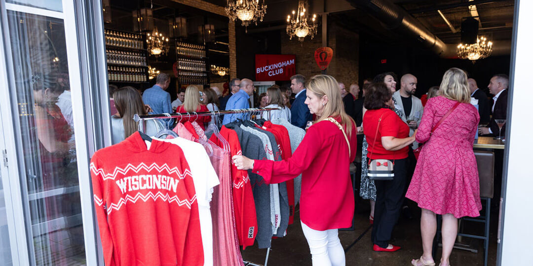 A group of people mingle at a social event with red and white "WISCONSIN" sweaters displayed on racks in the foreground; chandeliers and a "BUCKINGHAM CLUB" sign are visible in the background.