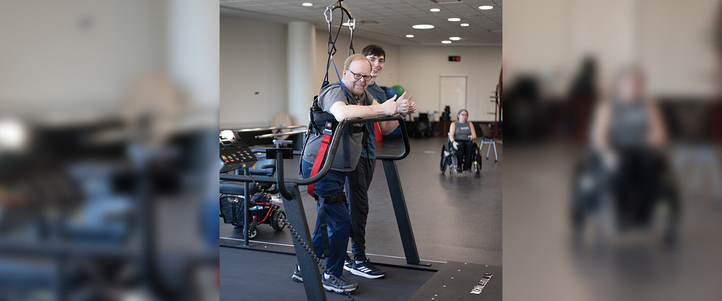 A man is standing on an in-ground treadmill and resting his arms on a support bar in front of him. He is supported by a black harness suspended from the ceiling. His arms are resting on a support bar in front of him, and he is looking at the camera and giving a double thumbs-up. Behind him, a young man walks with him on the treadmill and smiles at the camera.