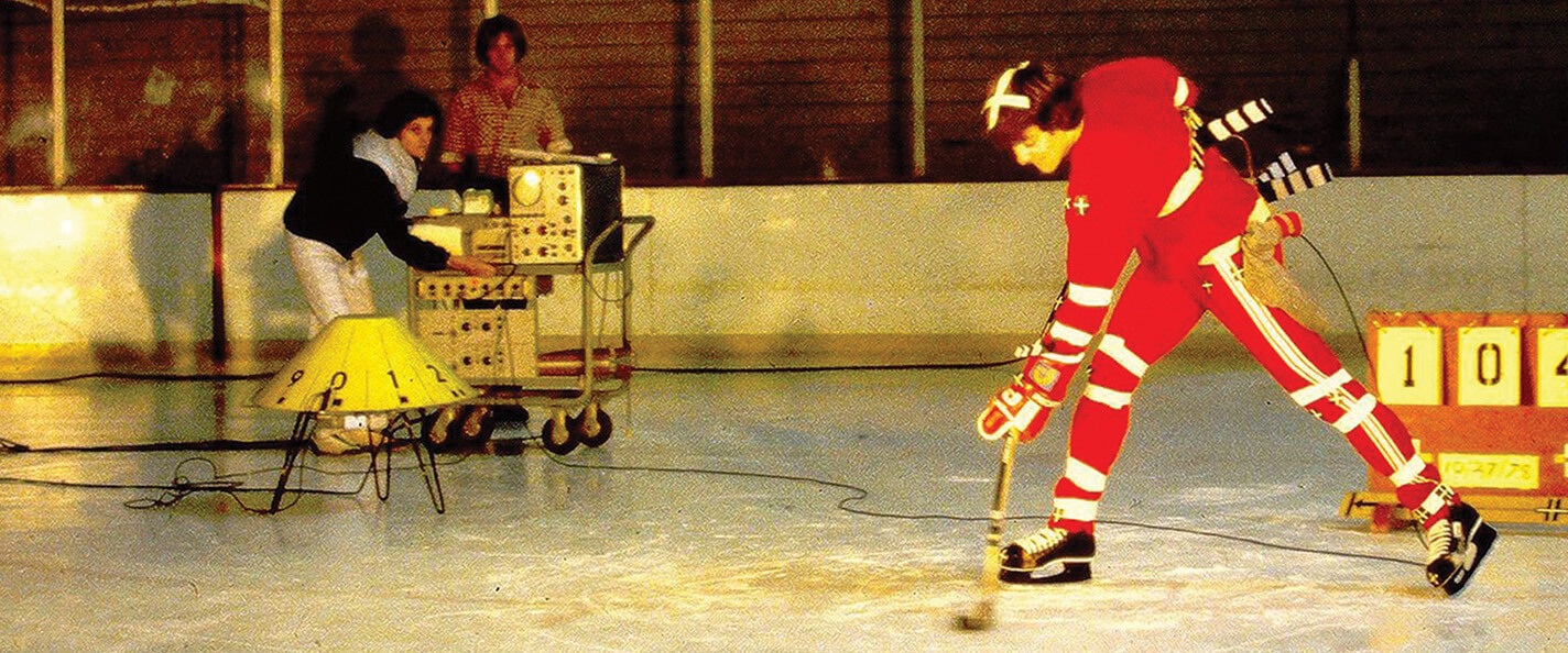 A study of hockey movement on an indoor ice rink. At back left, two researchers operate equipment while watching the study subject. At right (foreground), a hockey player in a red uniform with white accents wears kinetic tape and is attached to the equipment. He leans over in a position to shoot a puck.