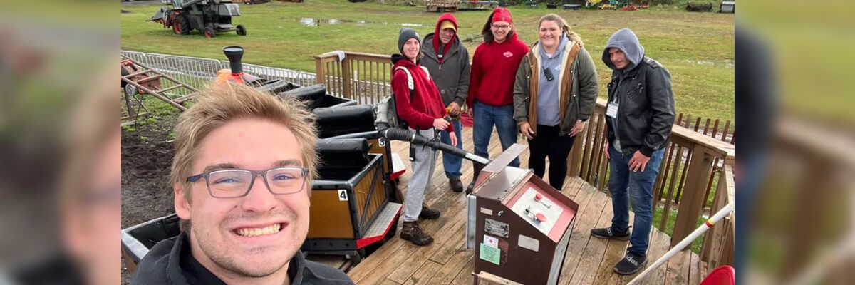 A young man with short blonde hair and glasses takes a selfie with a group of people. They are standing on a wooden platform next to a control panel for an amusement park ride.