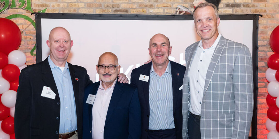 Panelists Kelly Sheffield, Sanjay Asthana, Jeff Wendorf, and Chris McIntosh stand in front of a backdrop with red and white balloons, dressed in formal attire.