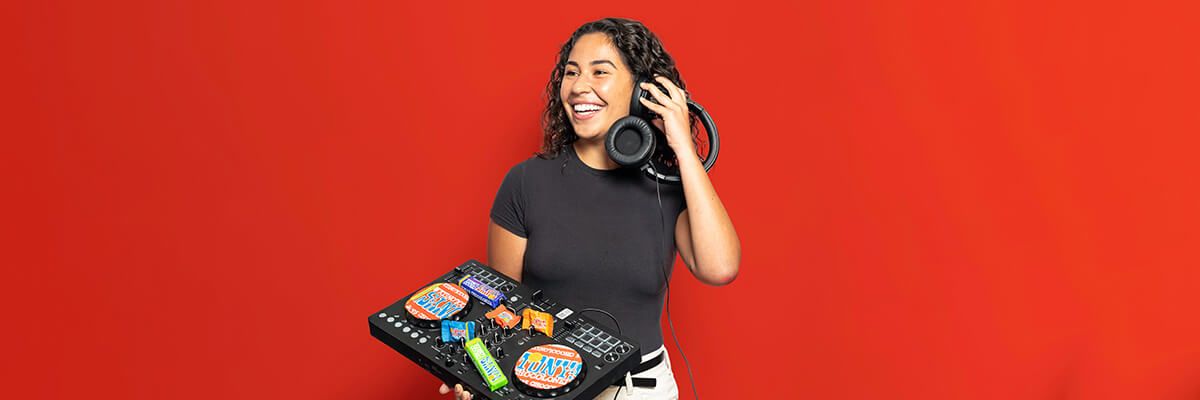 A woman with dark, shoulder-length, curly hair wearing a black T-shirt and white jeans stands against a red background. She holds one earpiece of a headphone set to her right ear and holds a small turntable with her left arm. The turntable is adorned with colorful candy wrappers. She is smiling and looking off to the left.
