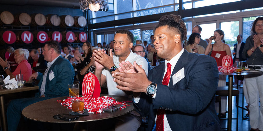 Attendees seated at tables clap during a social or professional event; tables are decorated with drinks and red items featuring the letter "W."