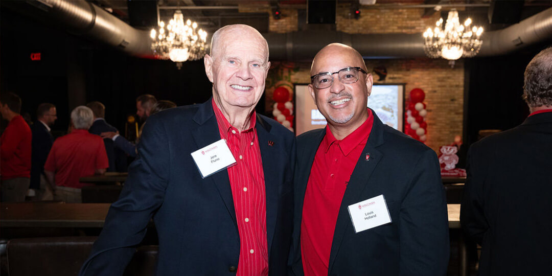 Jere Fluno and Louis Holland, both wearing dark blazers over red shirts with name tags, stand side by side at a social event with chandeliers and balloon decorations in the background.