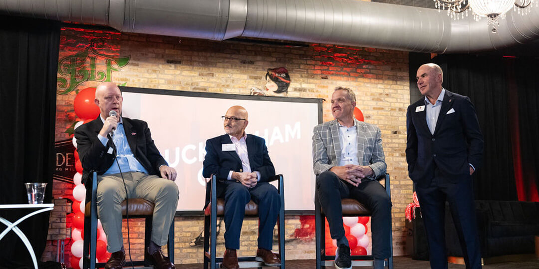 Panel discussion featuring Kelly Sheffield, Chris McIntosh, Sanjay Asthana, and Jeff Wendorf seated in front of a brick wall with artwork.