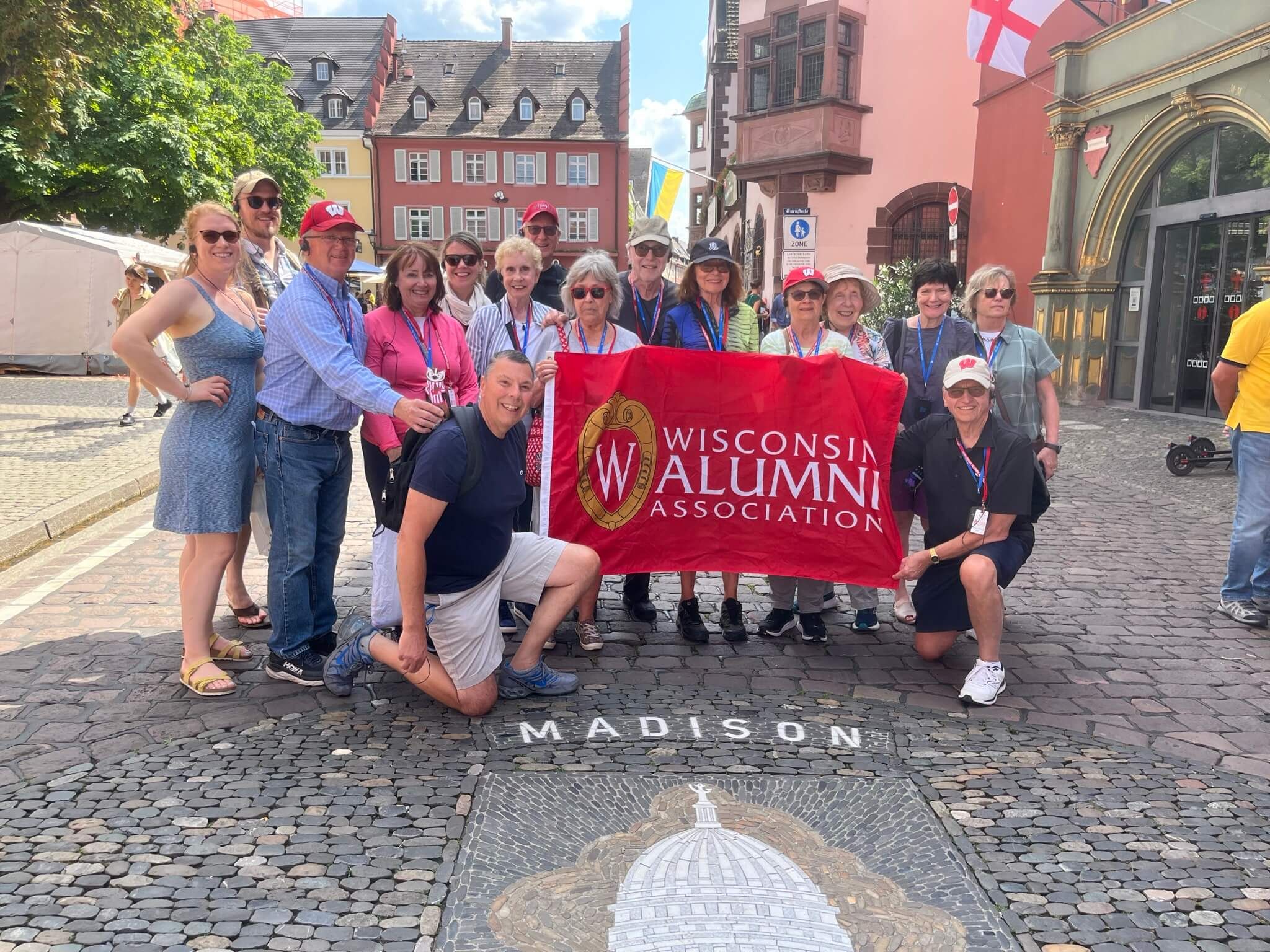 A group of tourists gather around a red flag that reads "Wisconsin Alumni Association" on a cobblestone plaza that features a mosaic of a white capitol dome under the word "Madison."
